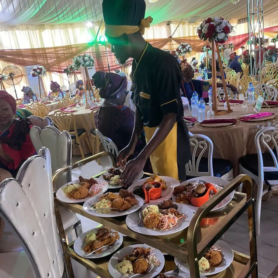 A steward plating jollof rice and asun from a service trolley inside a fully dressed marquee