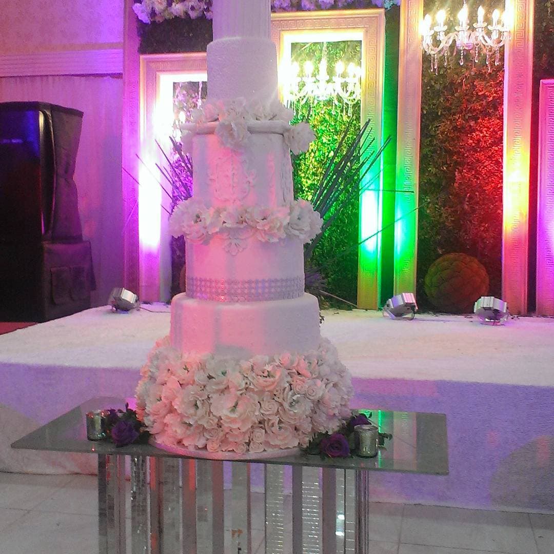 A five-tier white wedding cake ringed with sugar roses on a mirrored plinth under coloured uplighting