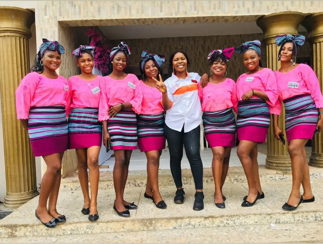 Eight Event Squad ushers in pink blouses and striped wrappers lined up on the steps of a venue portico