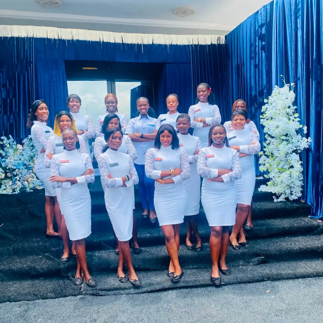 Fifteen ushers in white on a carpeted staircase in front of a blue velvet drape and white floral columns