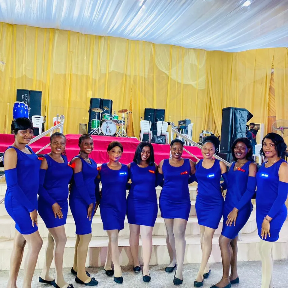 Nine ushers in royal blue standing in front of a dressed hall with the live band's stage behind them
